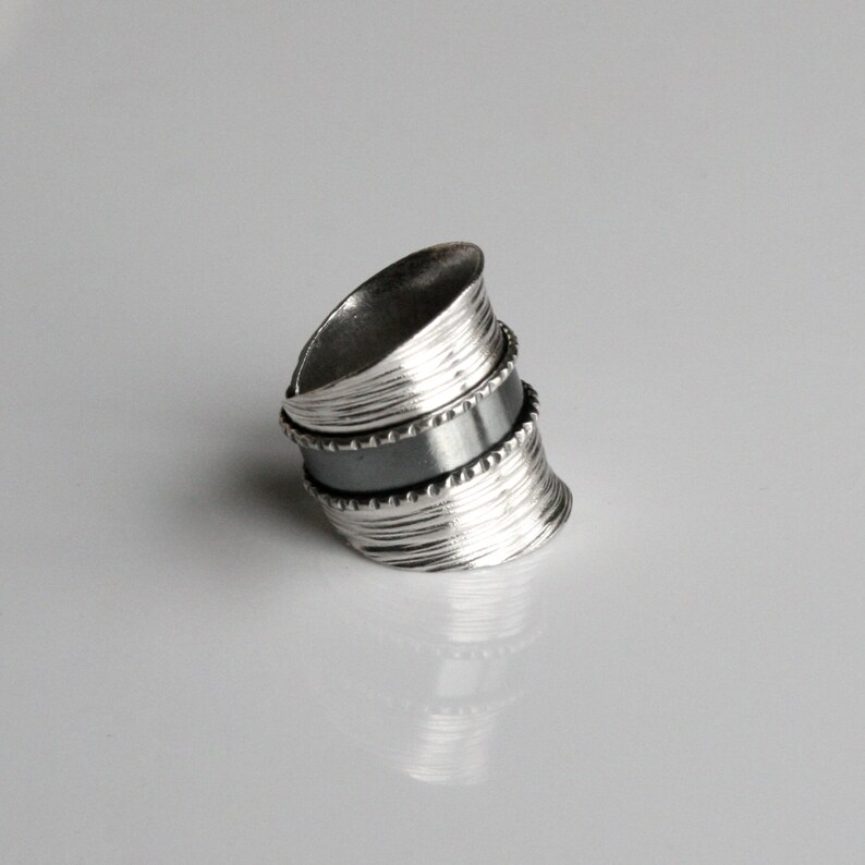 May include: A silver ring with a textured, layered design. The ring has a black band in the middle.