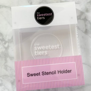 May include: A clear plastic stencil holder with a pink and white striped background. The text "Sweet Stencil Holder" is printed on the front of the holder. The logo "the sweetest tiers" is printed on the top of the holder.