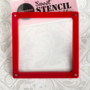 May include: Red plastic stencil frame with a clear plastic insert. The frame has a silver metal corner piece on each corner. The frame is on a white background with a floral pattern.