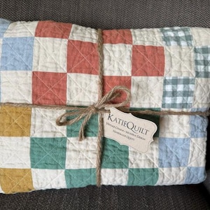 May include: A folded quilt with a patchwork design in shades of red, blue, green, and yellow squares, tied with twine. A tag with the text "KATIEQUILT" is attached. The quilt is a vintage charm, modern design, and handmade.