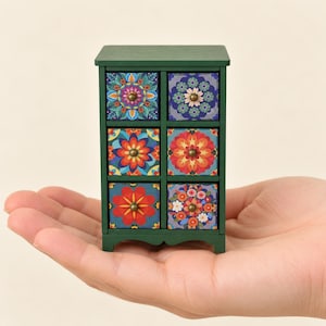 May include: A miniature dark green wooden chest of drawers featuring six individual drawers. Each drawer front displays a unique, colorful floral mandala pattern in shades of red, orange, blue, and yellow, finished with a small brass knob.