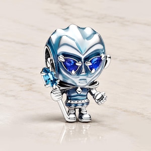 May include: A silver-toned charm shaped like a stylized character with a large head, deep blue gemstone eyes, and a blue-tinted body. The figure holds a staff topped with a light blue gemstone, featuring metallic blue accents and polished surfaces.