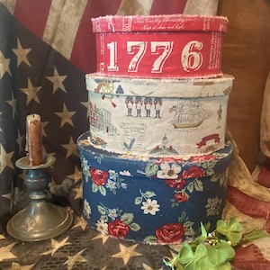 May include: A stack of three round fabric-covered boxes. The top box is red with the text 1776. The middle box features a cream background with illustrations of soldiers and ships. The bottom box is navy blue with a red and white floral pattern.