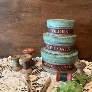 May include: A collection of vintage sewing supplies, including three stacked teal-colored round boxes with text that reads "COLORS", "J&P COATS", and "SPOOL COTTON". Wooden spools of thread, clothespins, and greenery are also present.