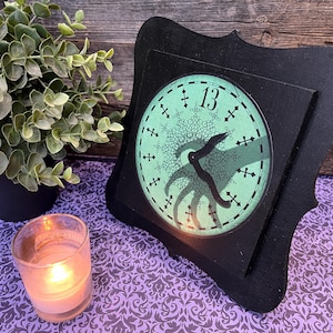 May include: A black framed clock with a teal face featuring the number 13 and a stylized tentacle design. A lit candle and a potted plant are in the foreground. The clock is on a purple patterned surface.