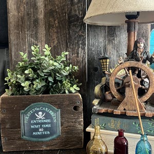 May include: A wooden box with a sign that reads "Pirates of the Caribbean Entrance Wait Time 80 Minutes". The box is decorated with a skull and crossbones. There are three small glass bottles on a wooden table in front of the box.