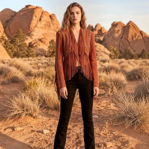 Puede incluir: Una chaqueta de ante color óxido con largos flecos en la parte delantera y las mangas, adornada con detalles de cuero trenzado y tachuelas metálicas, combinada con pantalones ajustados de color negro liso.