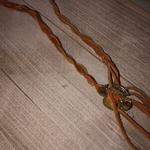 May include: A brown leather cord necklace with a gold metal charm. The charm is a braided design with a small bell attached.