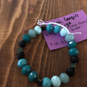 May include: A teal and light blue gemstone bracelet with black lava rock beads. The bracelet is tied with a white string and has a purple tag that says "Description:  Lava & Pitched Amethyst Bracelet - $14.18"