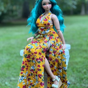 Puede incluir: Una muñeca con el pelo largo azul turquesa que lleva un vestido maxi amarillo con estampado floral. El vestido tiene un escote halter y una abertura alta en la parte delantera. La muñeca está sentada en un pedestal blanco.