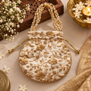 May include: A round, off-white drawstring pouch featuring intricate gold floral embroidery and sequin embellishments. The bag includes a handle made of multiple strands of small white faux pearls and gold-toned drawstring cords.