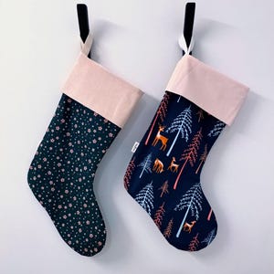 Peut inclure: Deux bas de Noël suspendus contre un mur blanc. L'un a un fond bleu foncé avec de petites fleurs roses. L'autre a un fond bleu foncé avec une scène de forêt, comprenant des cerfs et des arbres. Les deux ont un revers rose clair.