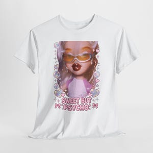 Può includere: Una t-shirt bianca a maniche corte con una stampa grafica di una bambola che indossa occhiali da sole color arancio. Il design include bolle, cuori, stelle e la scritta "SWEET BUT PSYCHO" in lettere rosa con piccoli fiocchi rosa ai lati.