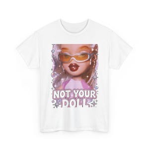 Può includere: Una t-shirt bianca con una stampa grafica di una bambola che indossa occhiali da sole color arancio e orecchini a cerchio. Il design include stelle scintillanti e bolle, con il testo NOT YOUR DOLL in un carattere grassetto e contornato.