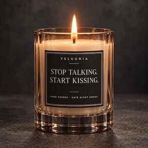 May include: A lit candle in a ribbed glass container with a black label featuring the text VELUUNIA STOP TALKING. START KISSING. HAND POURED DATE NIGHT SERIES. The wax is off-white and the flame is bright yellow with a soft orange glow.