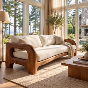 May include: A three-seat sofa featuring a curved, solid wood frame with a natural grain finish and thick, cream-colored fabric cushions. The sofa includes two matching throw pillows and a draped, light-colored woven throw blanket.