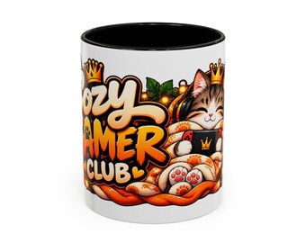 May include: A white ceramic mug with a black interior and rim, featuring a graphic of a tabby cat wearing a crown and headphones while holding a game controller, wrapped in a blanket with orange paw prints and the text Cozy Gamer Club.