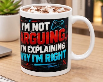 May include: A white ceramic mug filled with hot cocoa and marshmallows. The mug features a black background with a blue gaming controller icon and the text: I'M NOT ARGUING, I'M EXPLAINING WHY I'M RIGHT, printed in bold white, red, and blue lettering.