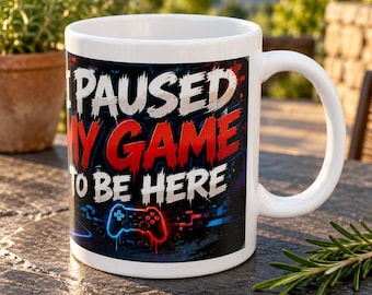 May include: A white ceramic mug featuring the text I PAUSED MY GAME TO BE HERE in bold white and red lettering. The design includes a graphic of a video game controller in blue and red against a dark, abstract, glitch-style background.