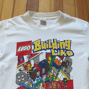 May include: White t-shirt with a colorful LEGO graphic. The graphic features a LEGO minifigure building with the text "Building Like Crazy".