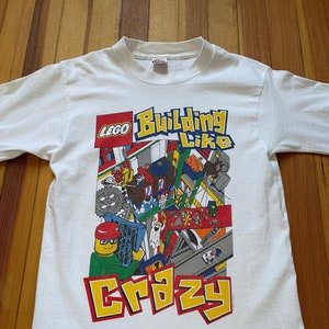 May include: White t-shirt with a colorful LEGO graphic. The graphic features a LEGO minifigure building with the text "LEGO Building Like Crazy".