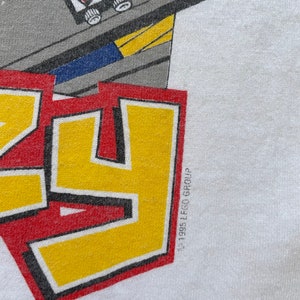 May include: A white t-shirt with a red, yellow, and black logo that says "LEGO" with a small LEGO man and truck in the background. The copyright symbol and "1995 LEGO Group" are printed in black on the bottom right corner of the shirt.