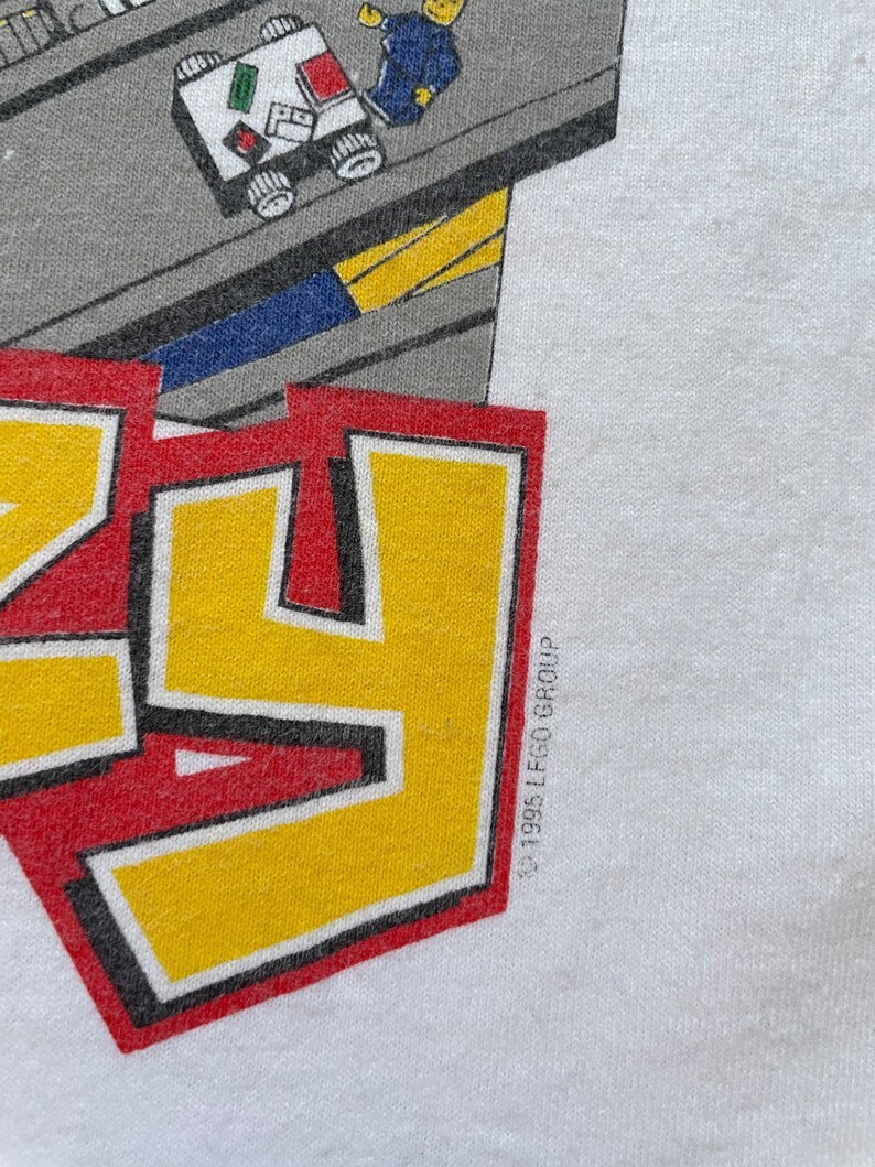 May include: A white t-shirt with a red, yellow, and black logo that says "LEGO" with a small LEGO man and truck in the background. The copyright symbol and "1995 LEGO Group" are printed in black on the bottom right corner of the shirt.