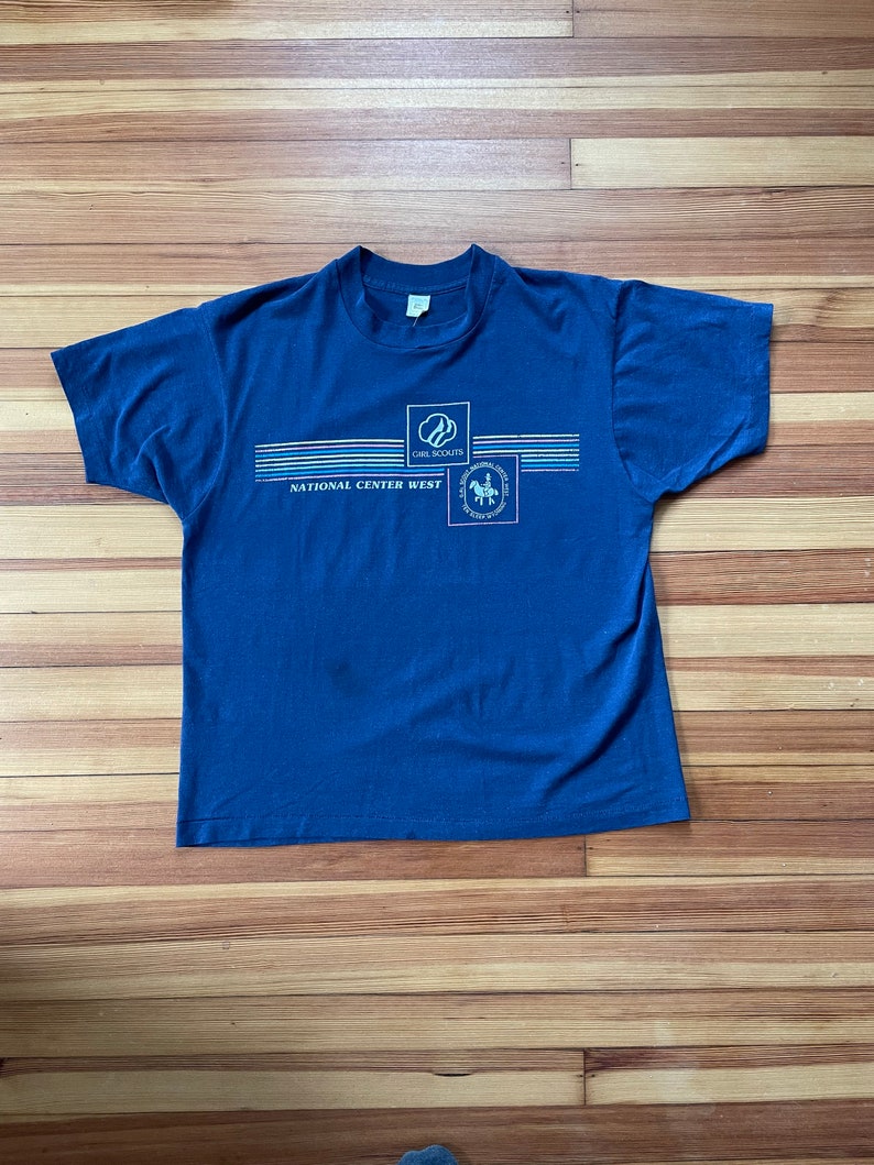 Puede incluir: Una camiseta azul marino con el texto "Girl Scouts National Center West" y un logotipo de Girl Scout.