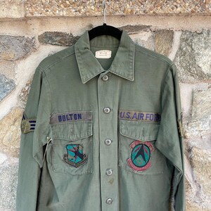 May include: A green military shirt with long sleeves and a button-up front. The shirt has two patch pockets on the chest and a name tag that reads "BOLTON". There are also two patches on the shirt, one with a blue and green design and the other with a red and green design. The shirt also has a patch on the left sleeve that reads "U.S. AIR FORCE".