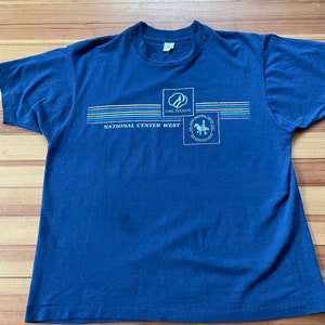 Puede incluir: Una camiseta azul marino con un dise&ntilde;o gr&aacute;fico que incluye el logotipo de Girl Scouts, las palabras "National Center West" y un dise&ntilde;o de rayas de colores.