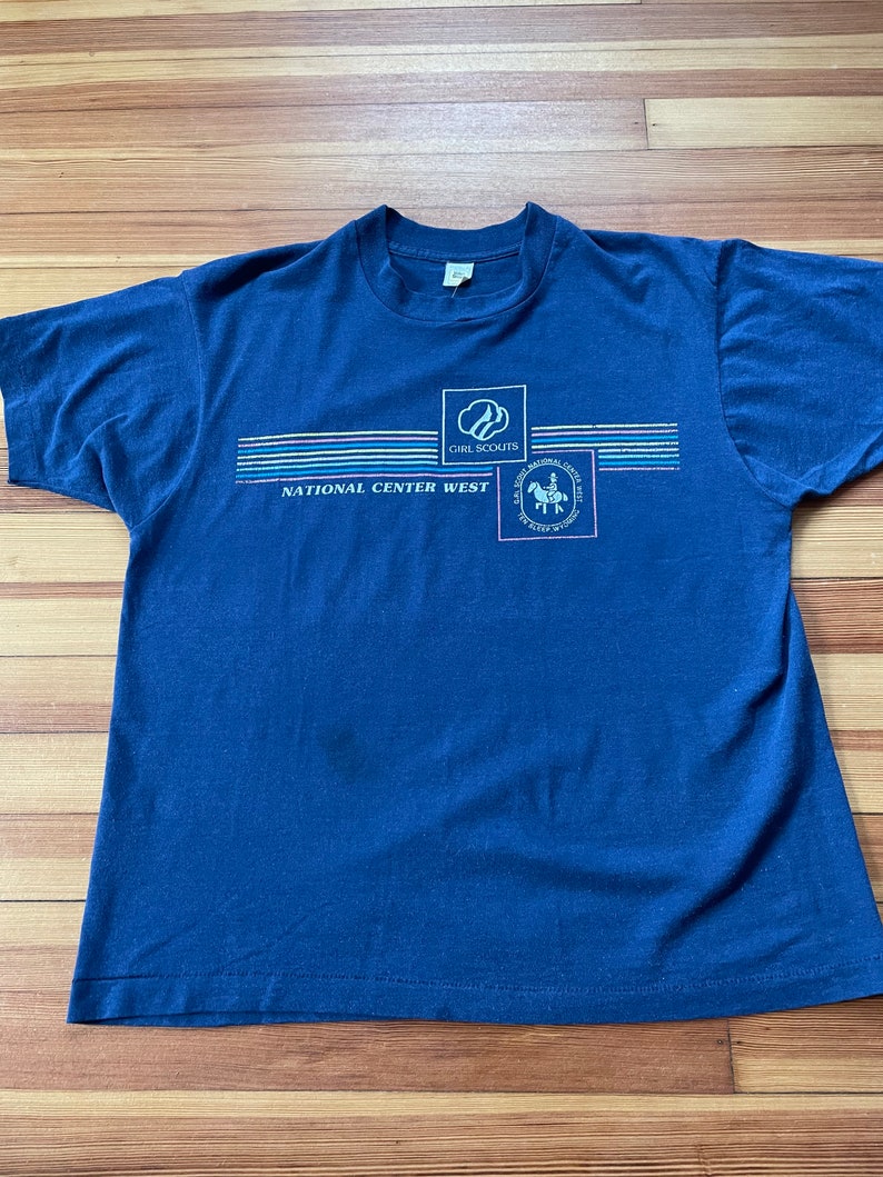 Puede incluir: Una camiseta azul marino con un dise&ntilde;o gr&aacute;fico que incluye el logotipo de Girl Scouts, las palabras "National Center West" y un dise&ntilde;o de rayas de colores.