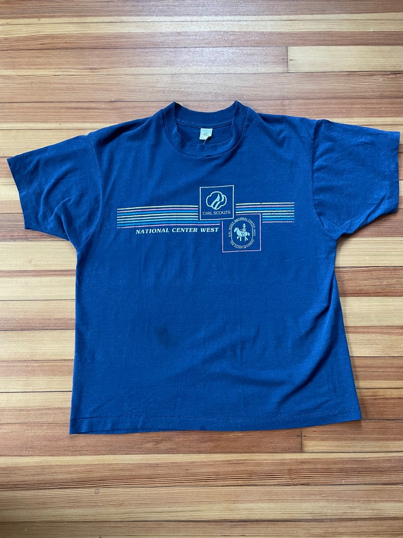 Puede incluir: Una camiseta azul marino con un logotipo de Girl Scouts National Center West. El logotipo presenta una silueta blanca de una ni&ntilde;a montando a caballo con las palabras "Girl Scouts National Center West" impresas debajo.