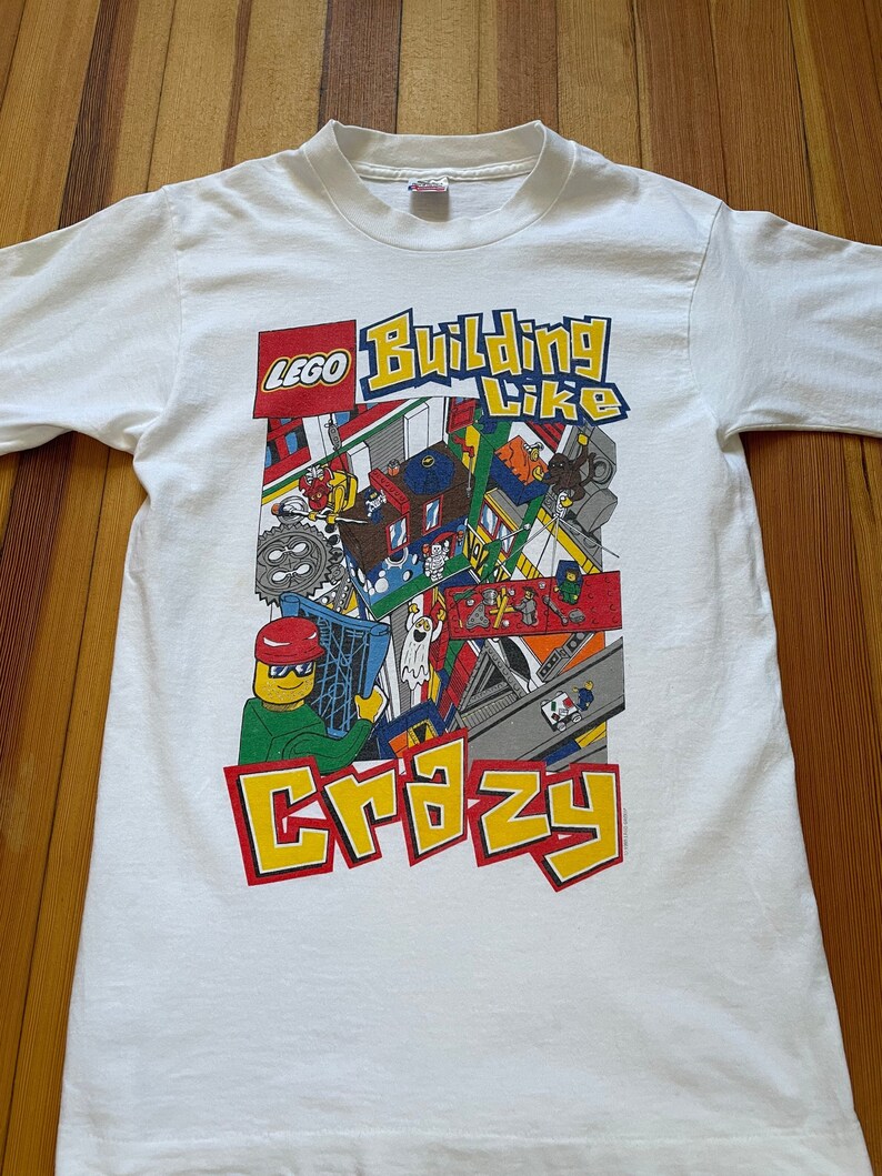 May include: White t-shirt with a colorful LEGO graphic. The graphic features a LEGO minifigure building with the text "LEGO Building Like Crazy".