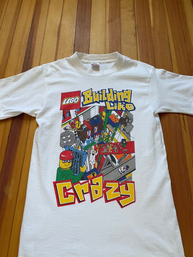 May include: White t-shirt with a colorful LEGO graphic. The graphic features a LEGO minifigure building with the text "Building Like Crazy".