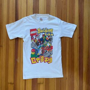 May include: White t-shirt with a colorful LEGO logo and the text "Building Like Crazy". The design features a cartoon illustration of LEGO bricks and minifigures.