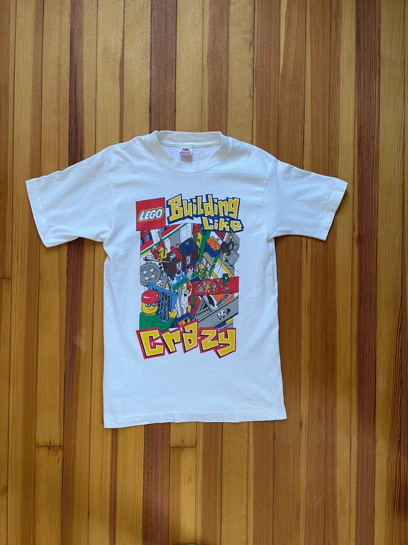 May include: White t-shirt with a colorful LEGO logo and the text "Building Like Crazy". The design features a cartoon illustration of LEGO bricks and minifigures.