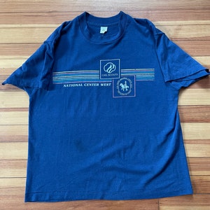 Puede incluir: Una camiseta azul marino con un logotipo de Girl Scouts y el texto "National Center West". El logotipo muestra a una ni&ntilde;a con uniforme de exploradora montando a caballo.
