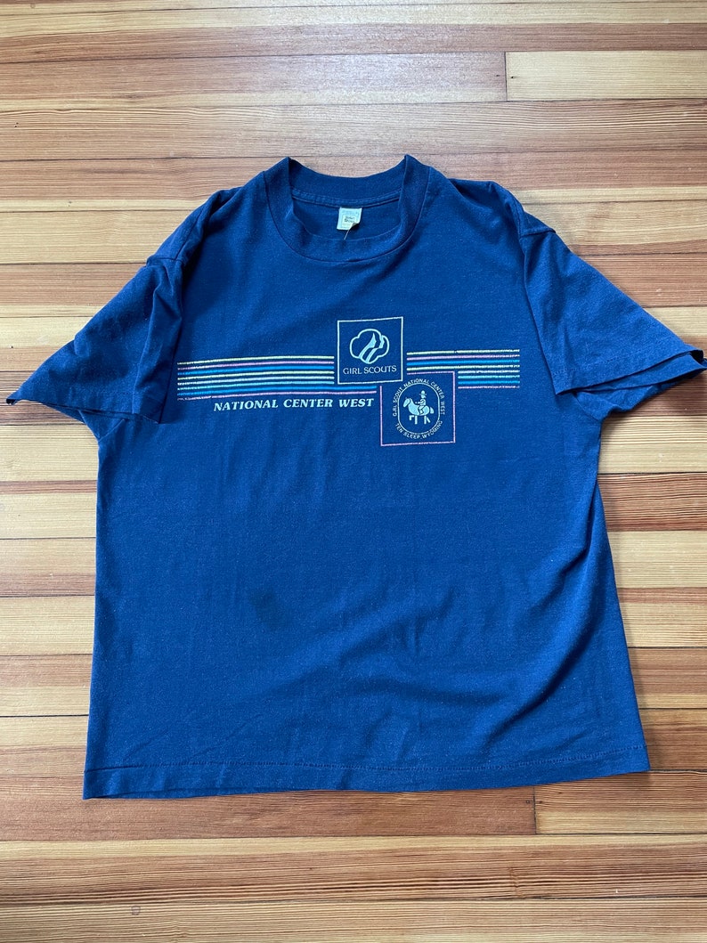Puede incluir: Una camiseta azul marino con un logotipo de Girl Scouts y el texto "National Center West". El logotipo muestra a una ni&ntilde;a con uniforme de exploradora montando a caballo.