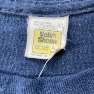 Puede incluir: Una etiqueta de camiseta azul con texto blanco que dice "50% Algod&oacute;n 50% Poli&eacute;ster Hecho en EE. UU." y "Velva Sheen Cincinnati Ohio, EE. UU." con una etiqueta de talla XL.