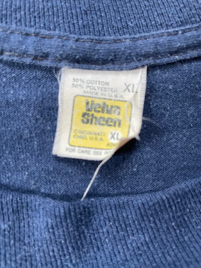 Puede incluir: Una etiqueta de camiseta azul con texto blanco que dice "50% Algod&oacute;n 50% Poli&eacute;ster Hecho en EE. UU." y "Velva Sheen Cincinnati Ohio, EE. UU." con una etiqueta de talla XL.