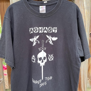 May include: A black short-sleeve t-shirt featuring a white graphic print. The design includes the text "AGHAST" at the top, two birds, a central sword piercing a skull, two circular symbols, and the text "SUMMER TOUR 2005" at the bottom.