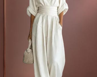 May include: A cream-colored jumpsuit featuring a deep V-neckline, wide three-quarter length sleeves with folded cuffs, and a wide, gathered waistband. The garment has loose, wide-leg trousers and includes a small, textured beige handbag.