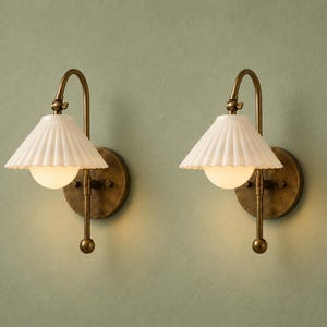 May include: A pair of wall sconces featuring a circular antique brass backplate, a curved arm, and a white pleated conical shade. Each fixture includes a central spherical light bulb that emits a warm glow, mounted on a vertical metal rod.