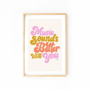 May include: A framed art print featuring the phrase "Music Sounds Better With You" in a retro style. The text uses pink, gold, and orange hues against a cream backdrop. The frame is a light wood colour.