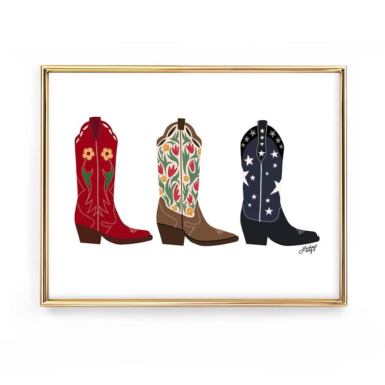 May include: A framed print featuring three colorful cowboy boots. The boots are red with yellow flowers, brown with orange and green floral patterns, and navy blue with white stars. The artwork is set against a white background.