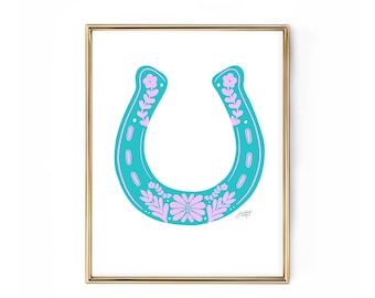 May include: A framed print featuring a turquoise horseshoe design with pink floral accents. The horseshoe is centered on a white background and framed in a gold-colored frame. The artwork has a folk art style.