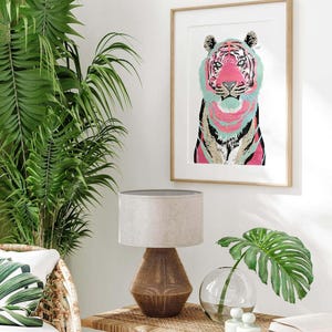 Puede incluir: Una impresi&oacute;n art&iacute;stica enmarcada con una colorida ilustraci&oacute;n de un tigre. El tigre tiene una cara rosa y rayas turquesas y rosas. La obra de arte se exhibe sobre una mesa con una l&aacute;mpara, un jarr&oacute;n y una hoja de monstera.