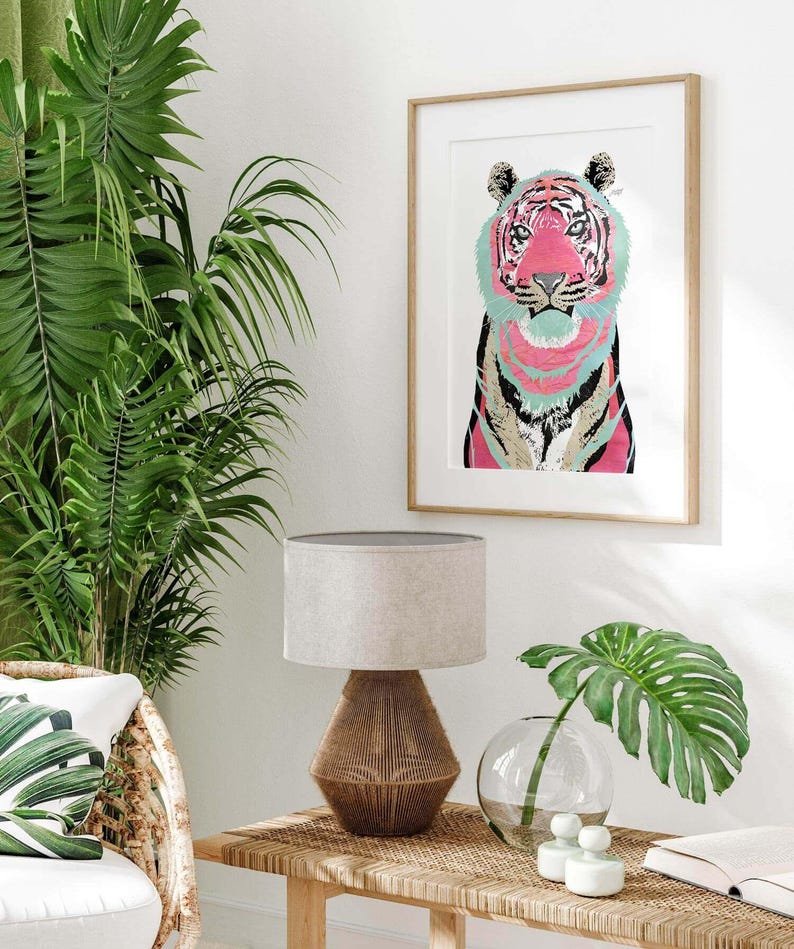 Puede incluir: Una impresi&oacute;n art&iacute;stica enmarcada con una colorida ilustraci&oacute;n de un tigre. El tigre tiene una cara rosa y rayas turquesas y rosas. La obra de arte se exhibe sobre una mesa con una l&aacute;mpara, un jarr&oacute;n y una hoja de monstera.