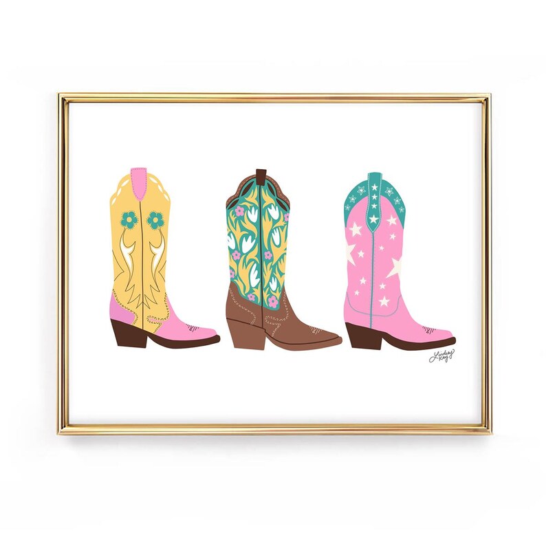 May include: A framed print featuring three colorful cowboy boots. The boots are in yellow, brown, and pink, each with unique floral and star designs. The boots have pointed toes and angled heels, set against a white background.