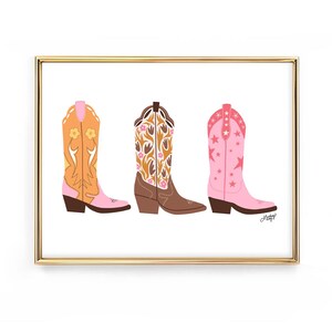 May include: A framed print featuring three colorful cowboy boots. The boots are in shades of orange, brown, and pink, each with unique floral or star patterns. The artwork is set against a white background.